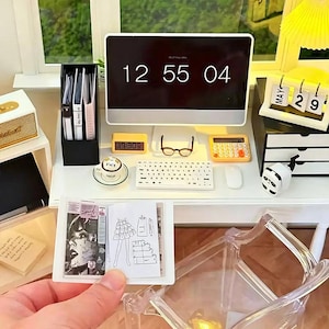 May include: Miniature desk scene with a computer displaying "12 55 04", a keyboard, calculator, and glasses. A small calendar shows "MAY 29". A hand holds a miniature magazine with a fashion sketch.