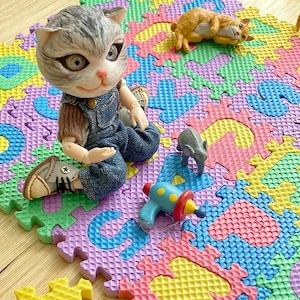 May include: A doll with a cat face wearing denim overalls and shoes sits on a colorful foam play mat. A toy ray gun, a small elephant figurine, and a toy cat are also on the mat. The mat is made of interlocking foam pieces.