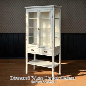 Puede incluir: Una vitrina alta de color blanco desgastado con dos puertas de cristal, tres estantes interiores, dos cajones pequeños con tiradores de latón y un estante inferior abierto. La vitrina mide 26,6 cm por 6,6 cm por 13,4 cm en total.