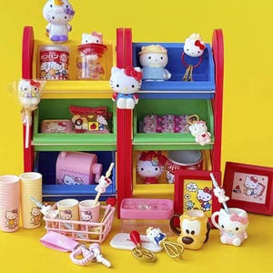Puede incluir: Una colorida exhibición de productos de Hello Kitty, que incluye figuras, tazas, un contenedor de almacenamiento rosa y una imagen enmarcada. Los artículos están dispuestos en un estante de varios niveles con secciones rojas, amarillas, verdes y azules, sobre un fondo amarillo brillante.