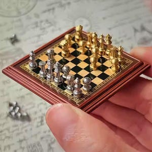May include: Miniature chess set with gold and silver pieces on a checkered board. The board is framed in dark wood with a decorative border. The chess pieces are arranged for a game, showcasing the intricate details of the tiny set.