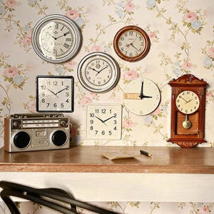 Peut inclure: Plusieurs horloges anciennes de formes variées, rondes, carrées et une horloge à pendule. Les horloges sont accrochées sur un fond de papier peint floral. Un boombox argenté est posé sur une surface en bois.