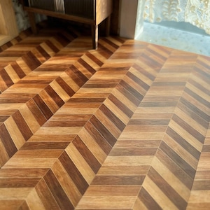 May include: A close-up shot of a wooden floor with a chevron pattern. The floor is made of alternating light and dark brown wood planks, creating a zig-zag design. The floor appears to be in a room with a wooden cabinet.