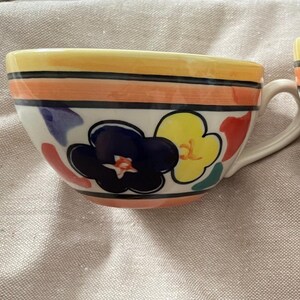 May include: Two ceramic cups featuring a hand-painted floral pattern with large dark blue and yellow flowers, accented by orange and black horizontal stripes. Each cup has a curved handle and a yellow rim, showcasing a colorful design.