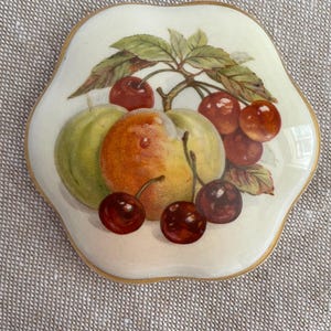 Può includere: Un piatto in ceramica color crema con bordi smerlati, caratterizzato da un'illustrazione centrale di ciliegie rosse, una mela verde e una pesca con foglie verdi. Il piatto presenta un sottile bordo color oro e una finitura lucida con crepe.