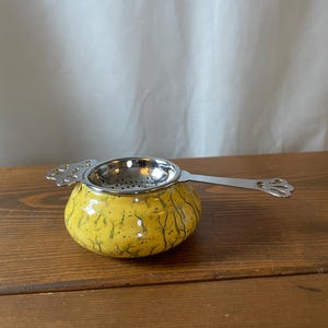 May include: A yellow tea strainer with a crackle glaze finish and a silver-toned metal rim. The strainer features a long, flat handle and a small side tab, both decorated with a cut-out floral pattern. The bowl of the strainer is perforated.