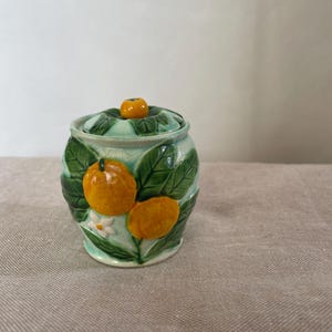 May include: A ceramic jar with a lid featuring a raised relief design of orange citrus fruits, green leaves, and a small white flower. The jar has a glossy finish with a light green background and a small orange fruit knob on the lid.