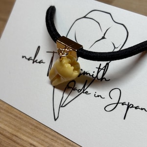 Op de afbeelding: Een zwarte haarband met een goudkleurige metalen sluiting en een kleine, crèmekleurige schelpenbedel. De bedel is met een kleine goudkleurige ring aan de sluiting bevestigd. De kaart achter de haarband heeft de tekst "Made in Japan".