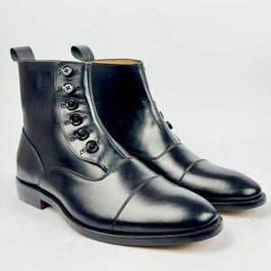 Könnte beinhalten: Ein Paar schwarze Lederstiefeletten mit Zehenkappe und einem seitlichen Verschluss mit sechs schwarzen Knöpfen. Die Stiefel haben eine glatte Oberfläche, eine strukturierte Silhouette und eine Zugschlaufe am oberen Fersenbereich zum Anziehen.