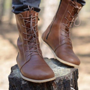 Könnte beinhalten: Ein Paar wadenhohe Stiefel aus braunem Leder mit einer Schnürung vorne, dünnen passenden Schnürsenkeln, einer abgerundeten Zehenpartie und einer flachen, hellen Sohle. Die Stiefel haben einen seitlichen Reißverschluss und sichtbare Nähte.