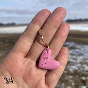 Puede incluir: Un colgante rosa en forma de corazón con motas oscuras cuelga de un lazo de metal dorado. El colgante se sostiene en una mano sobre un fondo borroso al aire libre. El texto "Simple Chaos Handcrafted by Michelle" es visible en la esquina inferior izquierda.