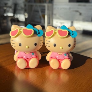 May include: Two Hello Kitty figurines. Each figurine is tan with black eyes and whiskers, wearing a pink swimsuit and red sunglasses. They have blue bows on their heads. The figurines are on a brown surface.