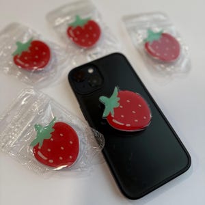 May include: A black smartphone with a red strawberry phone grip attached. Four additional strawberry phone grips are in clear plastic packaging. The strawberry grips have a red body, white seeds, and green leaves.