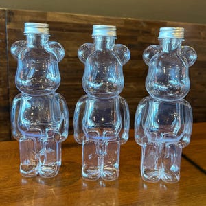 May include: Three clear plastic bear-shaped bottles with silver caps. The bottles are transparent, allowing a view of the contents. The bear design includes a head, torso, arms, and legs. The bottles are arranged in a row on a wooden surface.