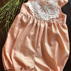 May include: A peach-colored baby onesie with a white crocheted lace overlay. The onesie has a gathered skirt and a ruffled neckline.