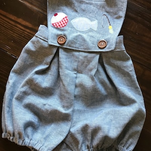 May include: A blue linen baby overall with a fishing theme. The overall has a white fish, a red and white bobber, and a fishing rod embroidered on the front. The overall has two brown buttons.