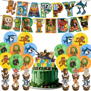 Puede incluir: Una escena festiva de fiesta de cumpleaños con una tarta verde, cupcakes, globos y una pancarta que dice "Happy Birthday". La tarta está cubierta con personajes de dibujos animados. Los globos presentan tiburones, ranas y otros personajes. Los cupcakes están decorados con varios adornos.