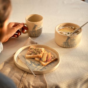 May include: A beige ceramic mug, bowl, and plate set with a blue and gray illustration of pumpkins and a fence. The plate holds slices of toast with toppings and apple slices. A spoon rests in the bowl of oatmeal.