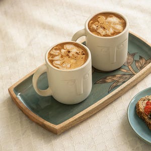 May include: Two cream-coloured ceramic mugs with a face design, filled with coffee and foam, sit on a wooden tray with a teal painted surface. The tray has a floral design and is next to a plate with a slice of bread.