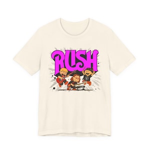 Può includere: T-shirt color crema con un'illustrazione di una band di cartoni animati. La parola "RUSH" è in rosa acceso. I membri della band suonano strumenti, tra cui una chitarra, una tastiera e una batteria. Linee nere si irradiano dai membri della band.