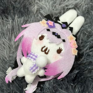 Puede incluir: Accesorio de peluche con forma de conejito en tonos lila claro y rosa. El conejito tiene orejas blancas, una cara adorable y está adornado con detalles florales y geométricos. Sostiene una pequeña estrella blanca con una bufanda a cuadros morada y blanca.