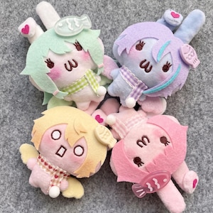 May include: Four plush bunny dolls in pastel colors: mint green, lavender, yellow, and pink. Each doll has a unique hairstyle, a scarf, and a small accessory. The dolls are lying on a gray surface.