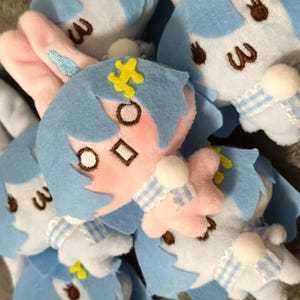 May include: A collection of plush bunny dolls. Each doll has a light blue head with a yellow flower, pink cheeks, and brown embroidered eyes. The dolls have white bodies with blue and white checkered bows and pink inner ears.