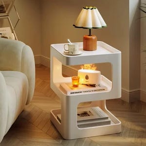 Könnte beinhalten: Ein weißer, moderner Beistelltisch mit integrierter Beleuchtung. Er verfügt über eine Lampe mit cremefarbenem Schirm, eine kleine Tasse und einen Diffusor. Der Tisch hat ein einzigartiges, offenes Design und steht neben einem Sofa.