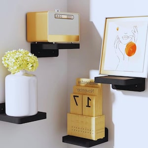 Puede incluir: Diseño interior moderno con estantes flotantes negros. Una estantería contiene un jarrón blanco con flores amarillas, otra una impresión artística enmarcada y un calendario de bloques de madera. Un electrodoméstico dorado y blanco está montado en una estantería.