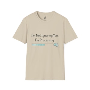 May include: A tan t-shirt with the text "I'm Not Ignoring You. I'm Processing." above a progress bar graphic and a brain illustration. The shirt has a crew neck and short sleeves.