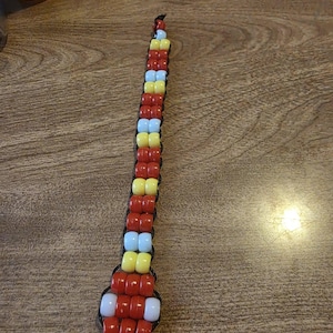 May include: A beaded lanyard featuring a snake design. The lanyard is made of red, yellow, light blue, and white beads strung on black cord. The snake's head is at the bottom, and a silver-colored ring is attached.