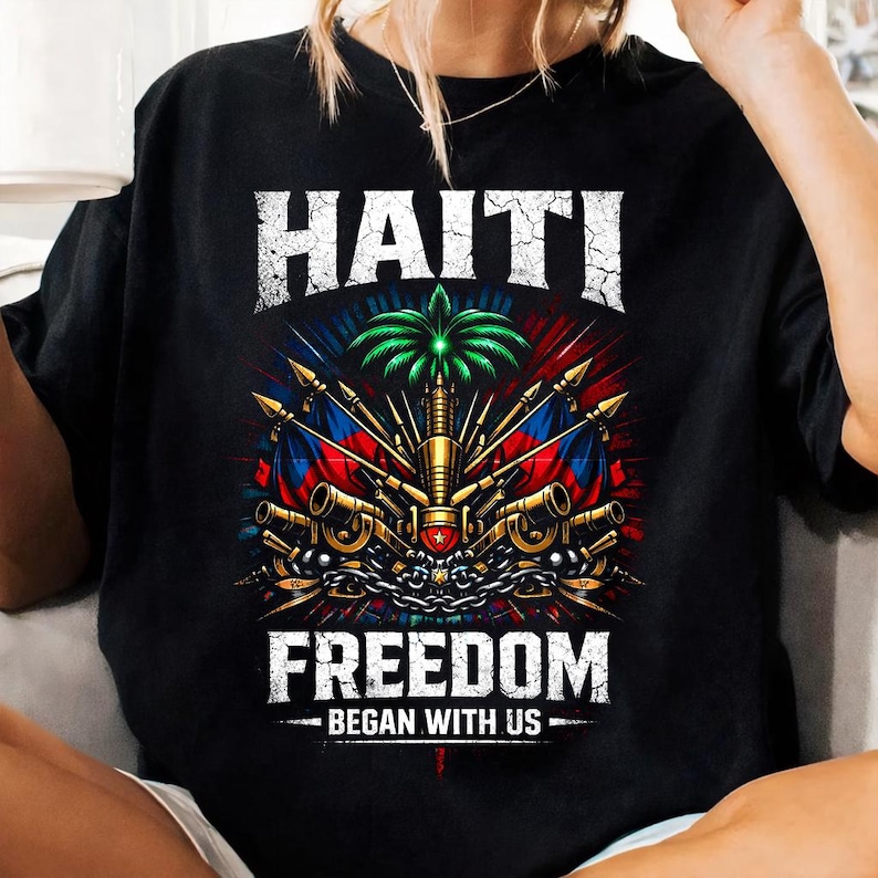 May include: Black t-shirt featuring a detailed graphic design with the word "HAITI" in white, above a central emblem with flags, palm tree, and weaponry. The text "FREEDOM BEGAN WITH US" is below the emblem.