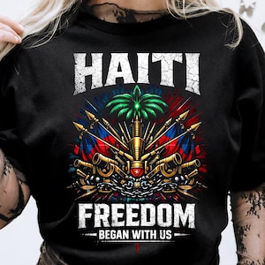 May include: Black t-shirt featuring a detailed graphic with the word "HAITI" above a central emblem. The emblem includes flags, cannons, and a palm tree, with the word "FREEDOM" below, and the phrase "BEGAN WITH US".