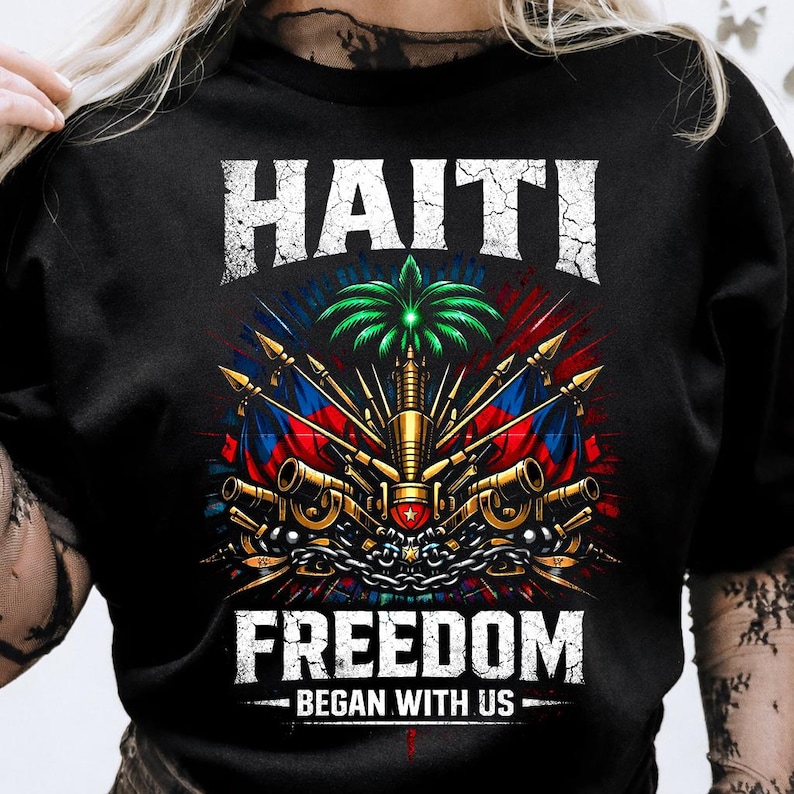 May include: Black t-shirt featuring a detailed graphic with the word "HAITI" above a central emblem. The emblem includes flags, cannons, and a palm tree, with the word "FREEDOM" below, and the phrase "BEGAN WITH US".
