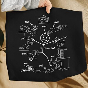 May include: Black tote bag with a white graphic depicting a stick figure surrounded by cats, with the text "LIFE WITH CATS" and "ow!" exclamations. The cats are shown in various playful and mischievous poses.