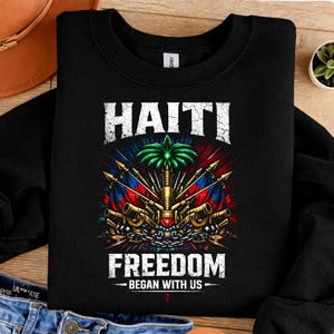 May include: Black sweatshirt featuring a graphic design with the word "HAITI" above a colorful emblem with flags, cannons, and a palm tree. The text "FREEDOM BEGAN WITH US" is below the emblem.