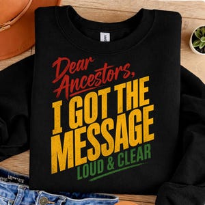 May include: Black sweatshirt featuring the text "Dear Ancestors, I GOT THE MESSAGE LOUD & CLEAR" in red, yellow, and green. The sweatshirt is made of a soft material and is suitable for casual wear.