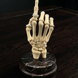 Puede incluir: Una escultura de mano esquelética de color hueso con el dedo medio levantado, que muestra tres anillos. La mano está montada sobre una base oscura y agrietada. Los anillos son plateados y dorados, uno de ellos con pequeñas piedras.