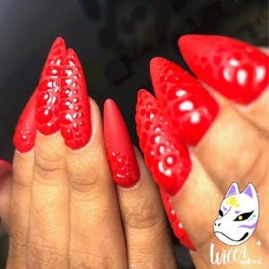 Peut inclure: Gros plan sur une main avec de longs ongles rouges en stiletto. Certains ongles présentent un motif texturé en forme de bulles. Les ongles sont brillants et vibrants. L'image comprend un logo de masque blanc avec des accents violets, jaunes et verts, et le texte "Wicca nail art."