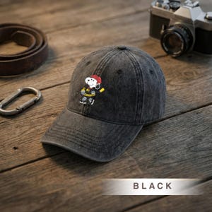 Peut inclure: Casquette de baseball noire avec un motif Snoopy joueur de hockey. La casquette a une visière incurvée et une finition vieillie. Le motif Snoopy comprend un casque rouge et un bâton de hockey jaune. Le mot "BLACK" est imprimé en bas.