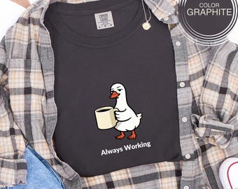 Op de afbeelding: Grafietkleurig t-shirt met een cartoon gans die een koffiekopje vasthoudt, met de tekst "Always Working" eronder. Het shirt is gelaagd met een geruit overhemd en een jeans.