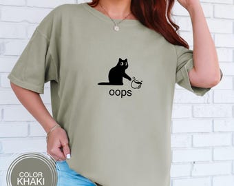 May include: Khaki-colored t-shirt featuring a black cat spilling a coffee cup, with the word "oops" below. The shirt has short sleeves and a relaxed fit. The color is indicated by a circular label that says "COLOR KHAKI".