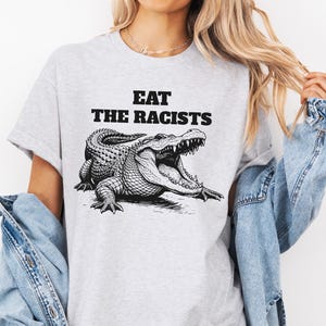 May include: A pale grey t-shirt with the words "EAT THE RACISTS" above a detailed black and white illustration of an alligator with its mouth open. The shirt is a casual, short-sleeved design.