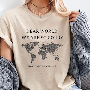 Pode incluir: Camiseta bege com a frase "DEAR WORLD, WE ARE SO SORRY" acima de um mapa-múndi preto. Abaixo do mapa, o texto diz "love, sane Americans". A camiseta tem mangas curtas e gola redonda.
