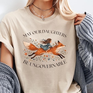 Puede incluir: Camiseta beige con una ilustración de una niña montada en un zorro, rodeada de flores. El texto dice "MAY OUR DAUGHTERS BE UNGOVERNABLE". La camiseta tiene mangas cortas y cuello redondo.