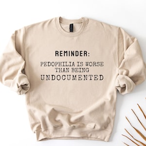 May include: A beige crewneck sweatshirt with the text "REMINDER: PEDOPHILIA IS WORSE THAN BEING UNDOCUMENTED" in black block letters. The sweatshirt has long sleeves and a ribbed neckline.