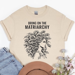 Puede incluir: Camiseta beige con el texto "BRING ON THE MATRIARCHY" sobre una ilustración en blanco y negro de Medusa. La ilustración muestra a Medusa con serpientes como pelo. Mangas enrolladas y cuello redondo.