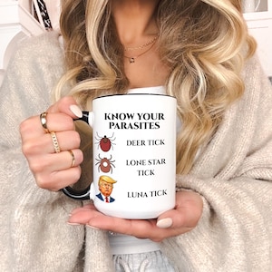 May include: White ceramic mug with a black handle and rim, featuring the text "KNOW YOUR PARASITES" and illustrations of ticks, along with a caricature of a politician. The mug is held by a person.