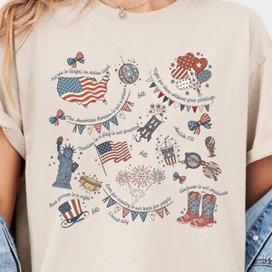 Op de afbeelding: Beige T-shirt met een patriottisch ontwerp. Het ontwerp bevat de Amerikaanse vlag, het Vrijheidsbeeld, vuurwerk en andere symbolen. Verschillende zinnen en citaten over vrijheid en rechten zijn gedrukt in rood, wit en blauw.