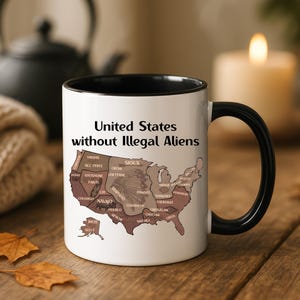 May include: A white ceramic mug with a black interior and handle. The mug features the text "United States without Illegal Aliens" above a map of the United States with tribal names. The mug is on a wooden surface with fall leaves.